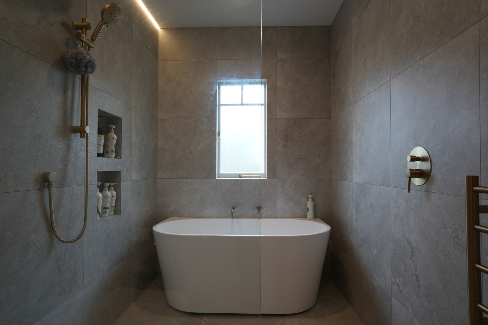 Freestanding bath suite Auckland — white oval bath in fully-tiled stone-look wetroom with brushed brass tapware